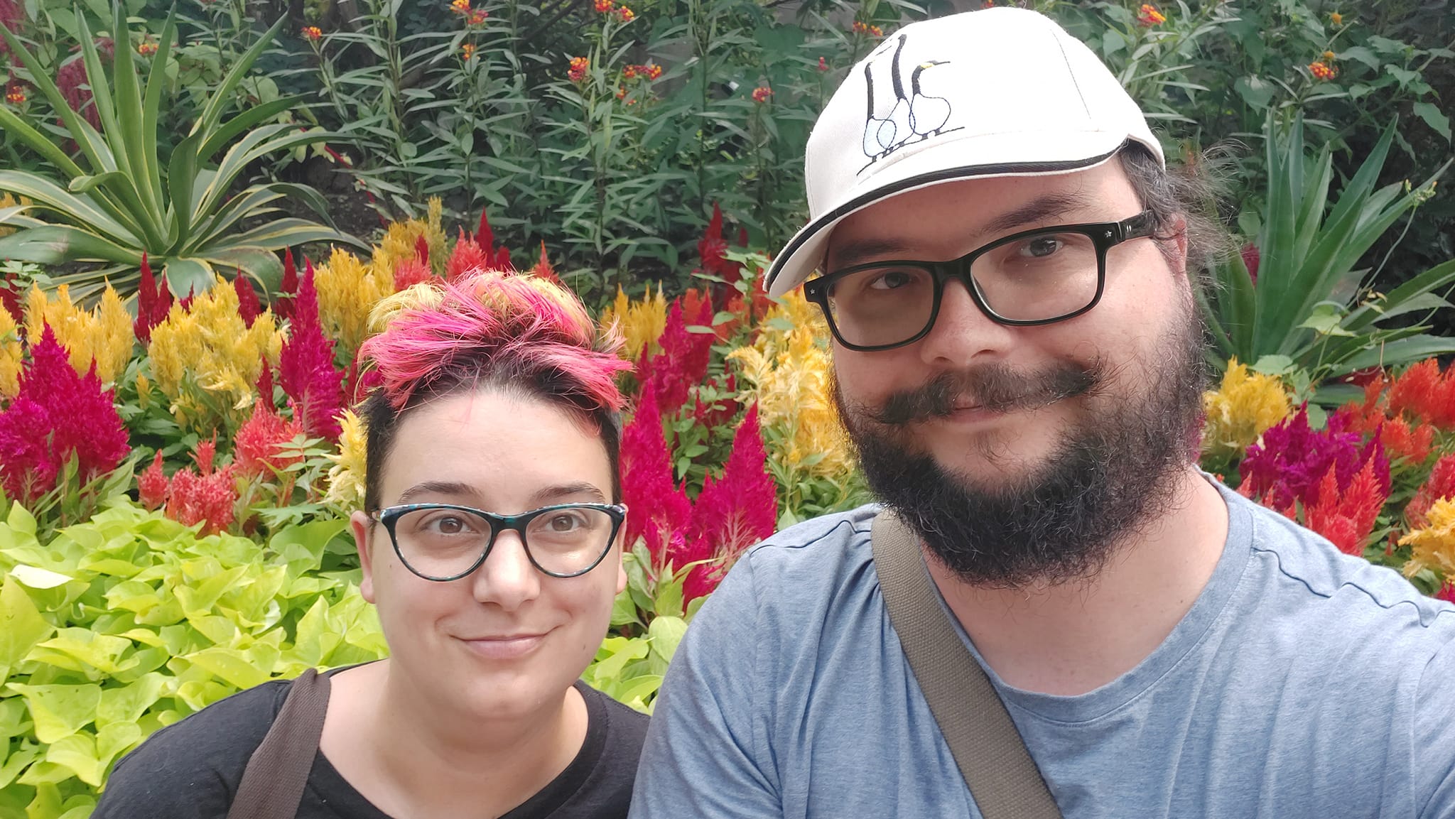 A man short hair and bushy facial hair sits next to a woman with a short rainbow mohawk in front of yellow and red flowers